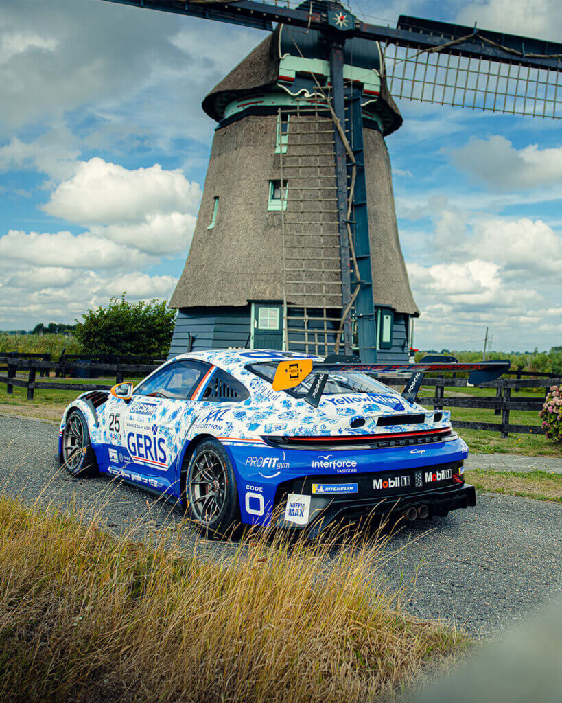 Blue-and-white race car with number 25 parked beside a thatched windmill in a rural setting.