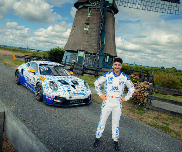 Race car with number 25 and sponsor logos parked beside a windmill; a driver in a white racing suit stands in front of it.